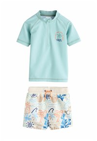 Una camiseta de protección solar verde menta con cremallera hasta la mitad, con un diseño de palmera. Acompañada de pantalones cortos de baño de color claro con un patrón tropical.