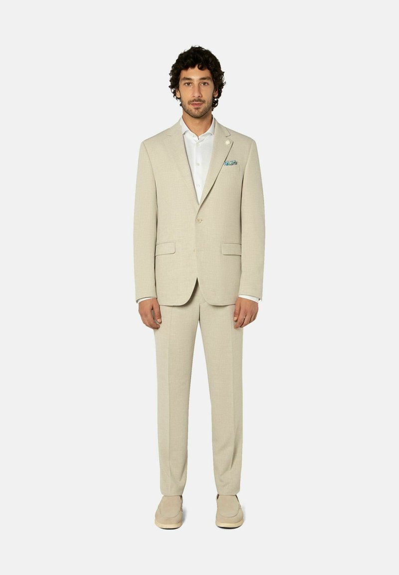 Mann stehend, trägt einen beigen Anzug mit weißem Hemd, gemustertem Einstecktuch und beigen Lederschuhen vor einem schlichten hellgrauen Hintergrund.