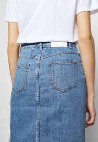 Person wearing a high-waisted blue denim skirt with back pockets and a white tucked-in short sleeve shirt, shown from the waist down.
