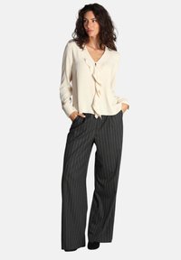 Blusa di crema chiaro con dettaglio arricciato sul davanti e maniche lunghe, abbinata a pantaloni larghi a righe sottili grigio scuro. Entrambi i tessuti appaiono lisci.