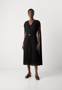 Black pleated dress featuring a V-neckline and an attached draped layer. A thin belt accentuates the waist. Paired with black slide sandals.