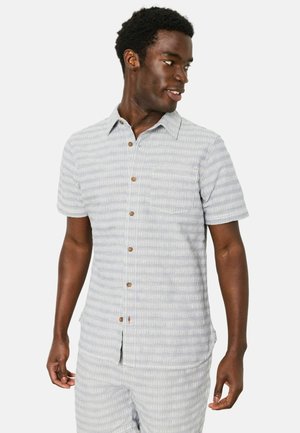 Mann trägt hellgraues und weiß gestreiftes Kurzarmhemd mit Knöpfen und passende Shorts, blickt nach rechts und lächelt.