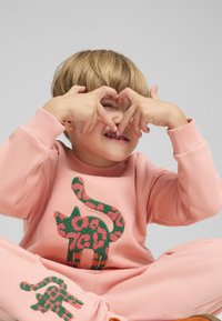 Conjunto de sudadera rosa con un diseño texturizado de gato en verde y rosa, que cuenta con un cuello redondo y puños acanalados. El niño forma un corazón con las manos.