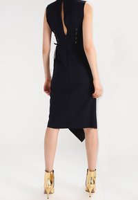 Marinblå ärmlös klänning med en djup rygcut, som har snörningsdetaljer och en hög-låg fåll. Gyldena ankelskor med en glänsande finish fulländar looken.