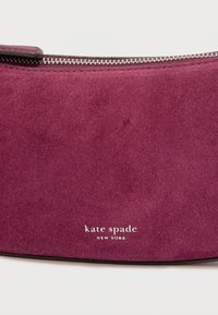 Sac à main en daim violet avec une fermeture éclair, portant le texte "kate spade NEW YORK" en blanc. Texture lisse, forme incurvée.