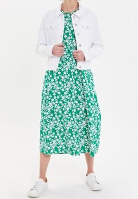 Chaqueta de mezclilla blanca sobre un vestido maxi floral verde con flores blancas; combinado con zapatillas blancas. La tela parece suave y ligera.