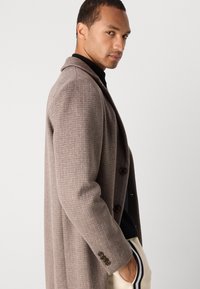 Abrigo largo marrón con un patrón y cuello, que presenta tres botones, llevado sobre un suéter oscuro. La vista lateral resalta el tejido texturizado.