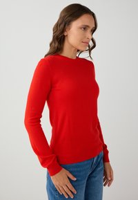 Pull rouge à col rond en tissu doux, avec des manches longues et un ourlet côtelé ajusté. Porté avec un jean bleu.