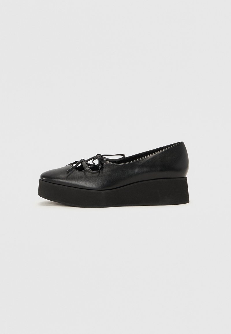Sapatos de plataforma em couro preto com bico arredondado, apresentando uma sola preta plana e um laço decorativo na parte frontal.