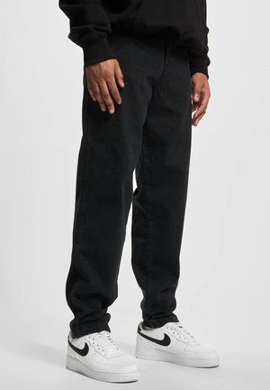 Homme portant un pantalon noir et des baskets blanches Nike avec des logos Swoosh noirs, debout sur un fond clair.