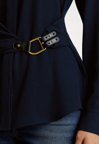 Nāvijuma blūze ar vērpjamu dizainu, melnu ādas sprādzi un zelta akcentiem, gluda tekstūra un apaļa apakša.