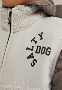 Veste matelassée en tissu crème avec un col texturé et duveteux. Texte "SALTY DOG" noir sur le devant et manches à motif camouflé.