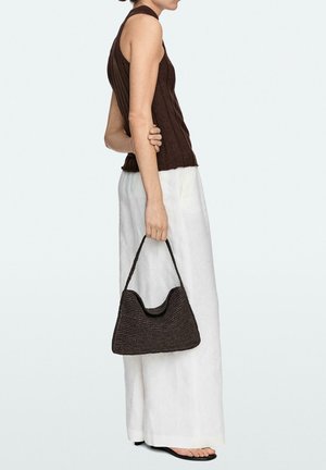 Frau in ärmellosem braunem Stricktop und weißen weiten Hosen, die eine dunkle gewebte Handtasche hält und im Profil vor hellem Hintergrund steht.