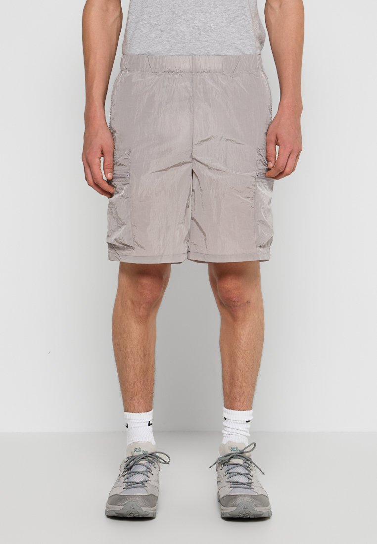 Lichte, geweven beige shorts met een elastische tailleband, zijvakjes en een gladde textuur, gecombineerd met grijze sportschoenen en witte sokken.