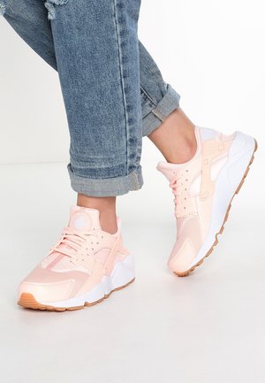 Osoba nosząca jasnoróżowe sportowe buty i podwinięte niebieskie dżinsy na jednolitym białym tle.