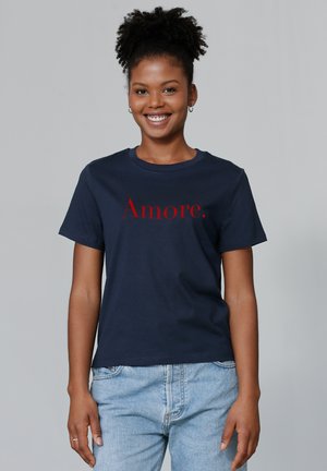 T-shirt en coton bleu marine à manches courtes, avec un graphique rouge "Amore." sur le devant. Design classique à col rond. Porte avec un jean délavé.