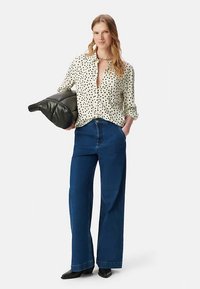 White shirt with black polka dots, fitted at the waist, paired with wide-leg blue jeans. Holds a black, padded clutch. Black shoes.
