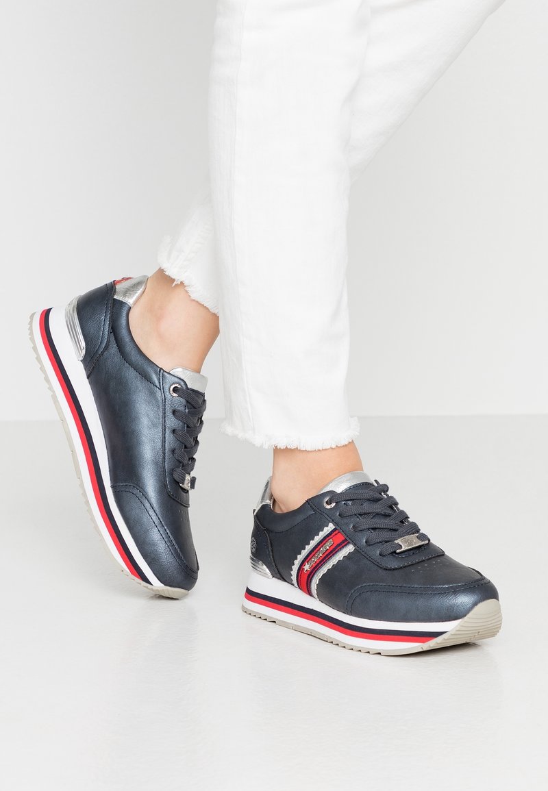 Personne portant des baskets bleu foncé avec des rayures rouges et blanches, associées à un pantalon blanc à ourlet effiloché, debout sur un sol blanc.