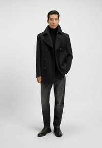 Cappotto doppiopetto nero con ampi revers, tessuto strutturato e bottoni neri; indossato sopra un dolcevita nero e abbinato a jeans scuri.