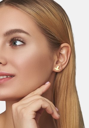 Gold bird-förmiger Steckerohrring am Ohr eines Models; glatte Textur, minimalistisches Design, kleine Größe, akzentuiert das Ohr, ohne überwältigend zu sein.
