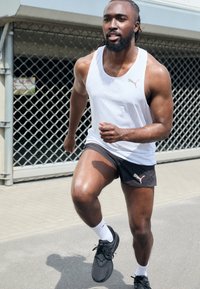 Top atletico bianco senza maniche con texture fine, pantaloni corti neri con logo. Scarpe da corsa nere, calze bianche. Posizione attiva sul marciapiede.