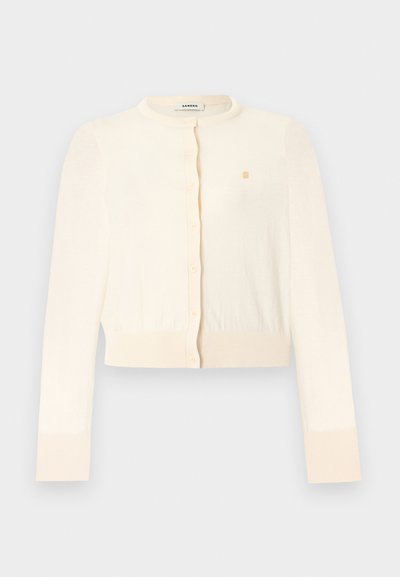 Cardigan court à manches longues crème avec encolure ronde, boutons sur le devant, poignets et ourlet côtelés, et petit logo doré brodé sur la poitrine.