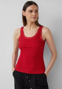 Rotes, geripptes Tanktop mit Spitzenbesatz am Rundhalsausschnitt und den Trägern. Das Kleidungsstück sitzt eng am Körper und wird mit schwarzen Hosen kombiniert.