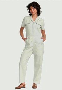 Woman with curly hair wearing light gray short-sleeve jumpsuit with front pockets and brown closed-toe sandals, hands in pockets, standing.
