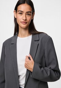 Szürke blézer sima anyagból, hajtókával és laza szabással. Fehér ing felett viselve, egyszerű, modern dizájnnal.