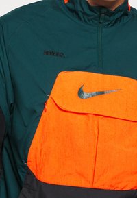 Chaqueta verde y negra con un bolsillo frontal de color naranja brillante que presenta un logo plateado de Nike y el texto "NIKE F.C." en el pecho.
