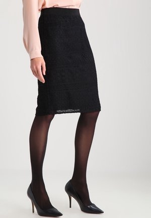 Woman wearing a black lace pencil skirt, sheer black tights, black high heels, and a light pink long-sleeve blouse, standing against a plain background.