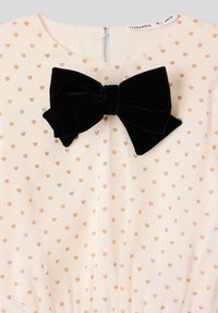 Abito di stoffa bianco con piccoli motivi a cuore dorato, caratterizzato da un grande fiocco in velluto nero al collo e una chiusura con zip sul retro.