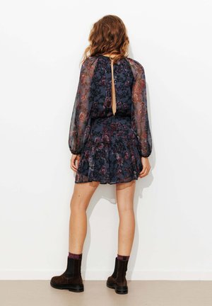 Femme aux cheveux ondulés portant une robe sombre à motif floral, courte, avec des manches longues transparentes, associée à des bottines marron et des chaussettes bordeaux.