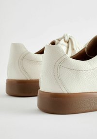 Scarpe in pelle bianca texture con suole in gomma marrone, dotate di semplici dettagli in pizzo e talloni arrotondati per un design elegante.