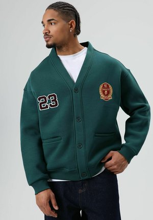 Groene, oversized cardigan met een V-hals, voorzien van twee voorkantzakken, zwarte knopen en geborduurd cijfer 23 en embleem op de borst.