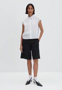 Camicia bianca a maniche corte con bottone abbinata a pantaloni corti neri fino al ginocchio e tacchi a punta neri, con calzini bianchi a scollatura.