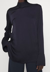 Blusa blu scuro a maniche lunghe con colletto alto, tessuto texturizzato e polsini larghi. Il taglio è morbido con orlo arrotondato.