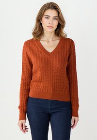 Pull en tricot couleur rouille avec un col en V, motif en relief de type torsade, manches longues et ourlet côtelé, associé à un jean bleu foncé.