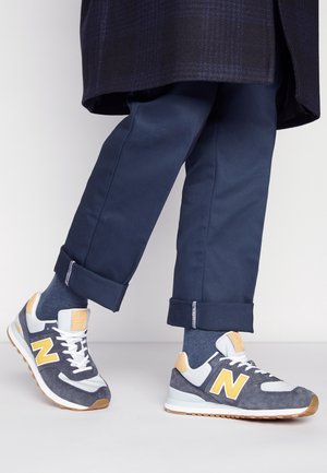 Personne portant un pantalon bleu marine à revers et des baskets New Balance grises, blanches et jaunes, debout sur un sol clair.