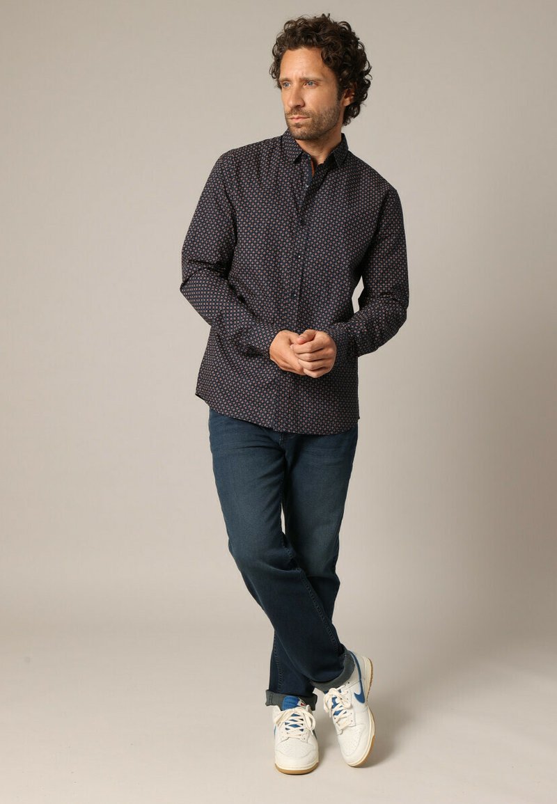 Donkergepatroneerd knoopshirt met kleine blauwe en rode motieven, gecombineerd met donkere denim jeans en witte sneakers met blauwe accenten.