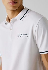 Biele polo tričko s jemnou textúrou, s navy pruhovaným golierom a logom Red Bull Racing na ľavej hrudi. Dve gombíky na zapínaní.