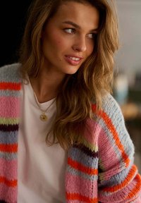 Mladá žena s dlouhými světlohnědými vlasy, hledící do strany, oblečená v bílé košili, barevném pruhovaném pleteném kardiganu a s kulatým přívěskem na náhrdelníku.