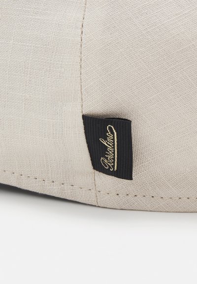 Beige tekstilpose med en strukturert overflate, utstyrt med en svart stofflabel med gullfarget kursivskrift. Sømdetaljer synlige på kanten.
