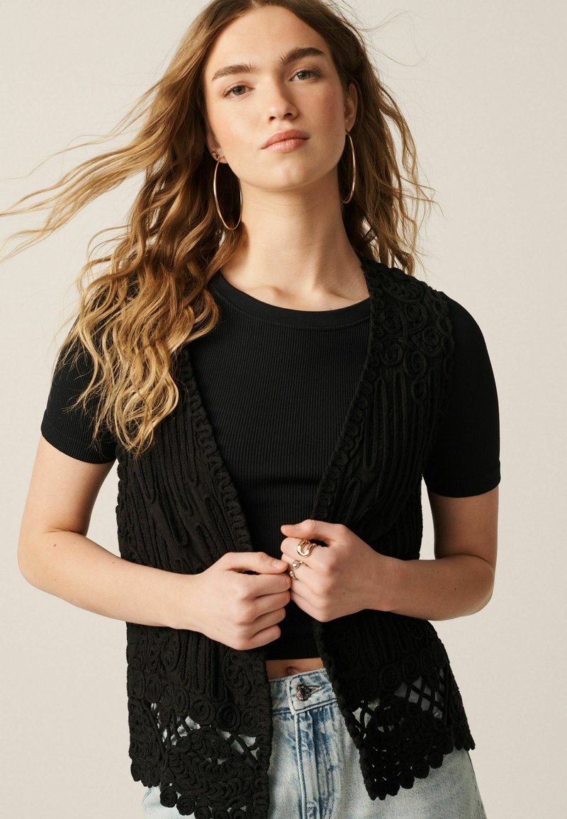 Vesta croșetată neagră cu modele intricate și design cu lucru deschis, purtată peste un top negru cu fante. Modelul poartă blugi și accesorii.
