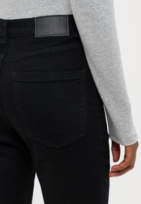 Jeans de denim preto com um design ajustado, apresentando um rótulo de couro na cintura e um bolso traseiro visível. Tecido texturizado.