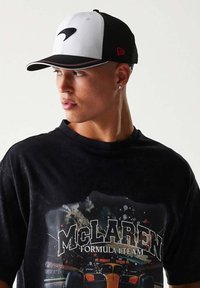 Czarny i biały kapelusz z zakrzywionym daszkiem. Posiada wyrazisty logo z przodu, czerwone akcenty i teksturowany materiał. Noszony z t-shirtem z grafiką.