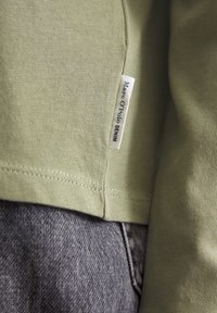 Olivengrønn bomullsskjorte med sydd hem, kombinert med grå denim. Merket viser "Marc O'Polo DENIM" i svart på en hvit tag.