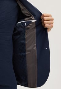 Mano che tiene aperto un blazer blu scuro mostrando la fodera interna con tessuto fantasia e l'etichetta "extra slim fit".
