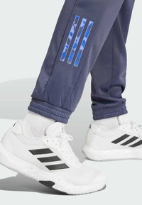 Pantalones deportivos de color navy con acentos de camuflaje azul en el dobladillo, combinados con zapatillas blancas que tienen rayas negras y un tejido de malla transpirable.