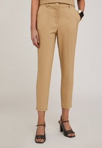 Pantaloni beige su misura con vestibilità slim, caratterizzati da una texture liscia, tasche laterali e un design frontale piatto. Abbinati a sandali neri con tacco.
